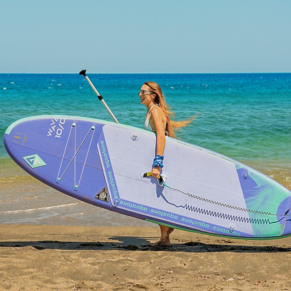 Mujer en la playa llevando el SUP Aquatone Wave 10’0 y su remo